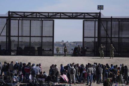 Migrantes y solicitantes de asilo que cruzaron la frontera de México a Estados Unidos esperan junto a un muro