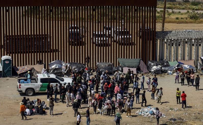 Migrantes y solicitantes de asilo que cruzaron la frontera de México a Estados Unidos esperan junto a un muro